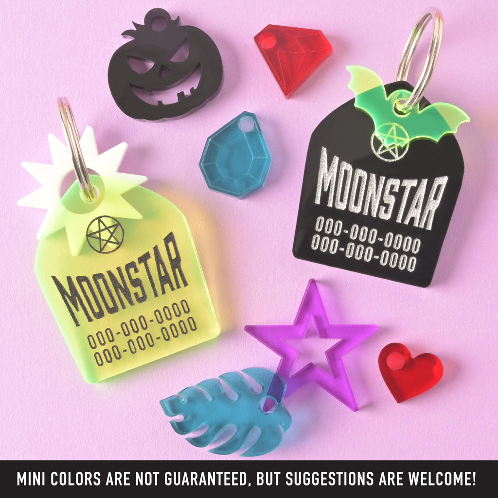 Two Gravestone personalized pet tags with a selection of mini charms scattered around on a solid lavender background. banner reads "mini colors are not guaranteed, but suggestions are welcome"