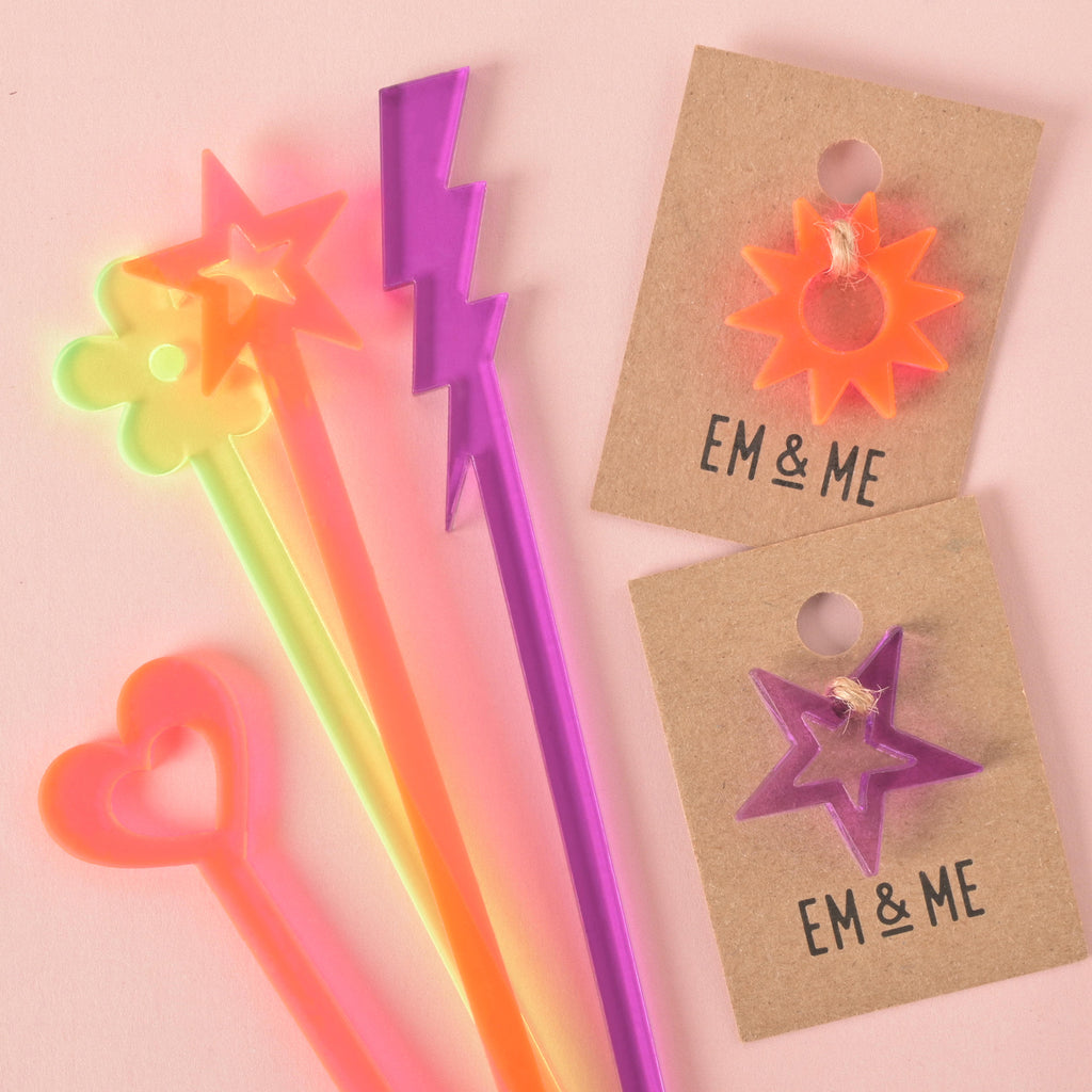 A collection of colorful stirrers, including a neon pink heart, a neon yellow flower, a Fluorescent pink star, and a purple lightning bolt. Next to the stirrers are two small Mii Charms—a neon pink sun and a purple star—on brown cards. The background is a solid pink color.