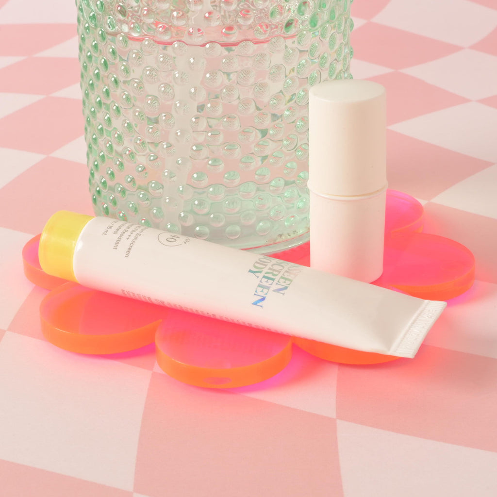 A textured, light green glass bottle and two sunscreen products—one in a tube and one in a stick—are sitting on a vibrant Fluorescent Pink flower-shaped coaster. The background is a pink and white checkered pattern.
