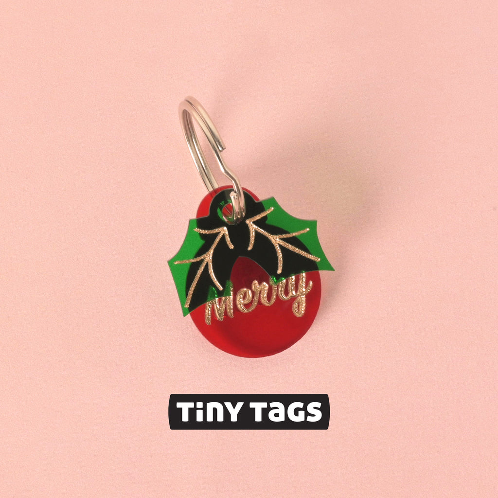 Close-up of a small, red circular pet ID tag with the glitter gold slogan 'Merry.' A green and black acrylic holly leaf charm is layered over the top. The tag is branded "TINY TAGS" and is displayed on a pink background.