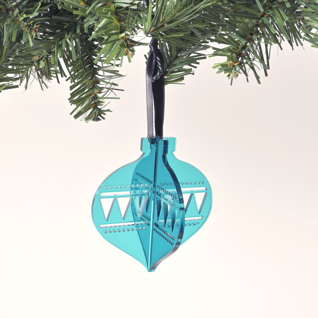 A teal, flat-pack Christmas tree ornament shaped like a classic glass ornament. The assembled ornament is hanging from a pine branch with a dark black ribbon. The ornament has a zig-zag and dotted design etched on the sides. The background is a solid white color.