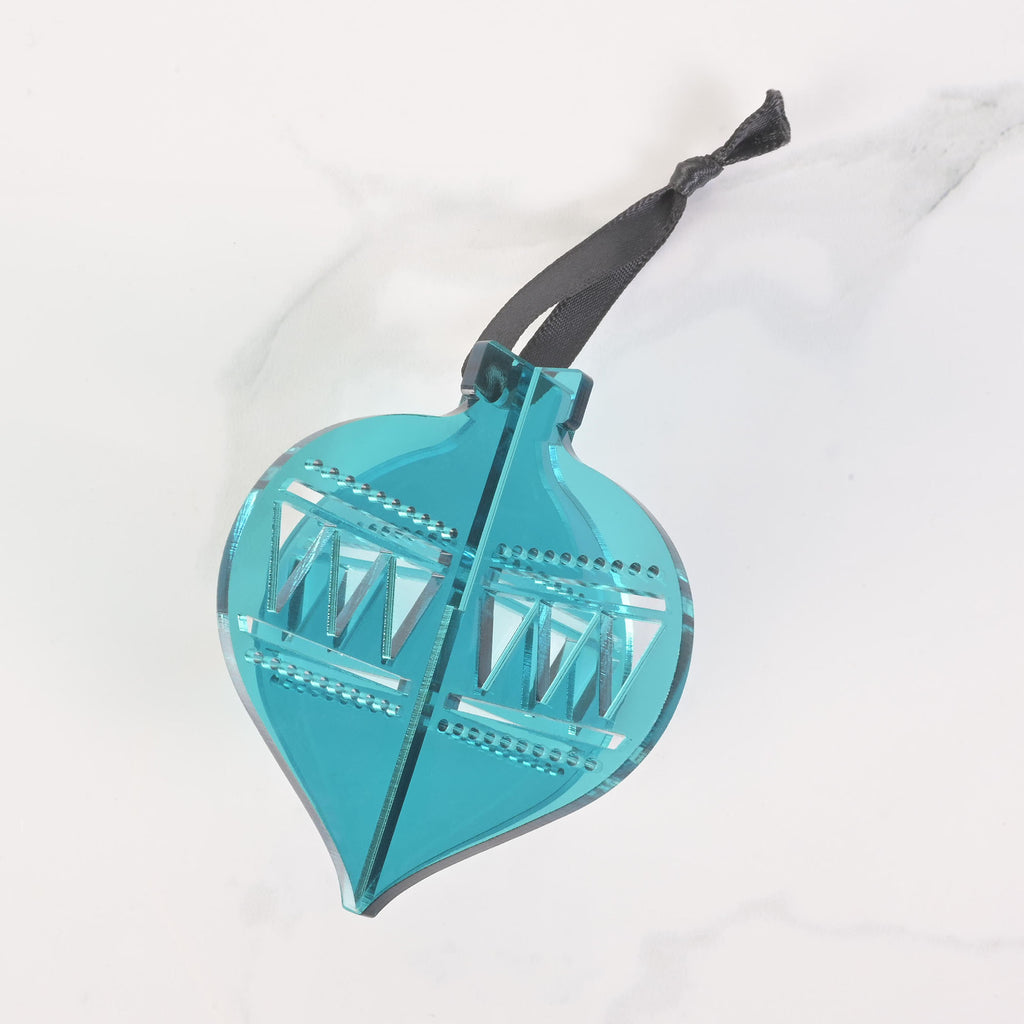 A teal flat-pack Christmas tree ornament shaped like a classic glass ornament. The ornament is assembled and has a dark black ribbon tied to the top for hanging. The item is displayed on a white marble surface.