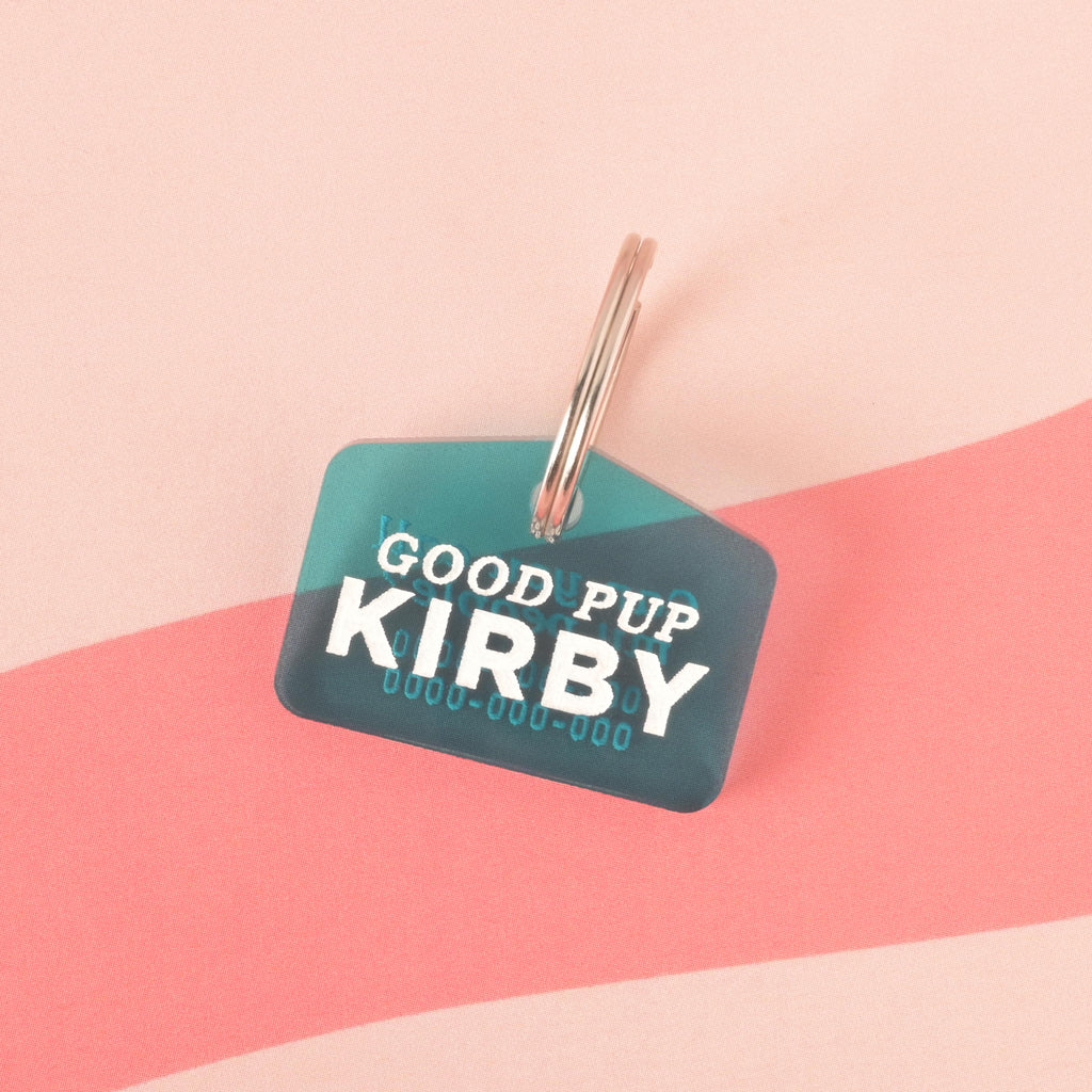 A personalized blue-green dog tag that says "GOOD PUP KIRBY" and has a placeholder phone number. The tag is attached to a silver ring, perfect for a collar, and is displayed on a pink background with a wavy design.