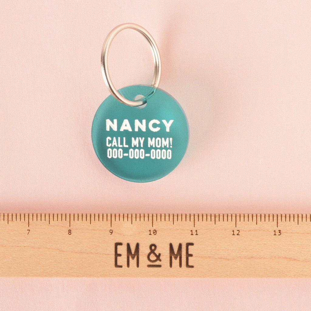 A close-up product photo of a round, teal pet tag, also known as a Minimal Circle tag. The tag is engraved with the name "NANCY" and the funny phrase "CALL MY MOM!" It is shown with a silver split key ring, and a wooden ruler with the words "EM & ME" is positioned below to show its size.