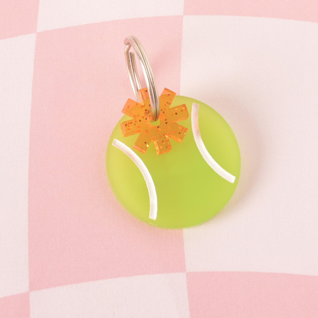 Close-up of a circular, neon yellow-green acrylic pet ID tag designed like a tennis ball. An orange glitter acrylic charm shaped like an asterisk/starburst is layered on the tag, attached to the silver split ring. The tag is displayed on a pink and white checkered background. Flower charm, Orange Asterisk Mini, Geometric Flower Seasonal Mini, Tiny Accessory, Add-on Charm