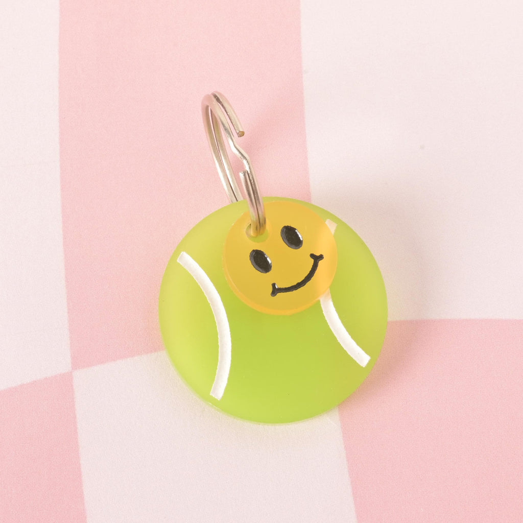 A custom, translucent green, circular pet ID tag shaped like a tennis ball with white lines. A small, yellow smiley face charm is attached to the silver keyring. The tags are displayed on a pink and white checkered background.