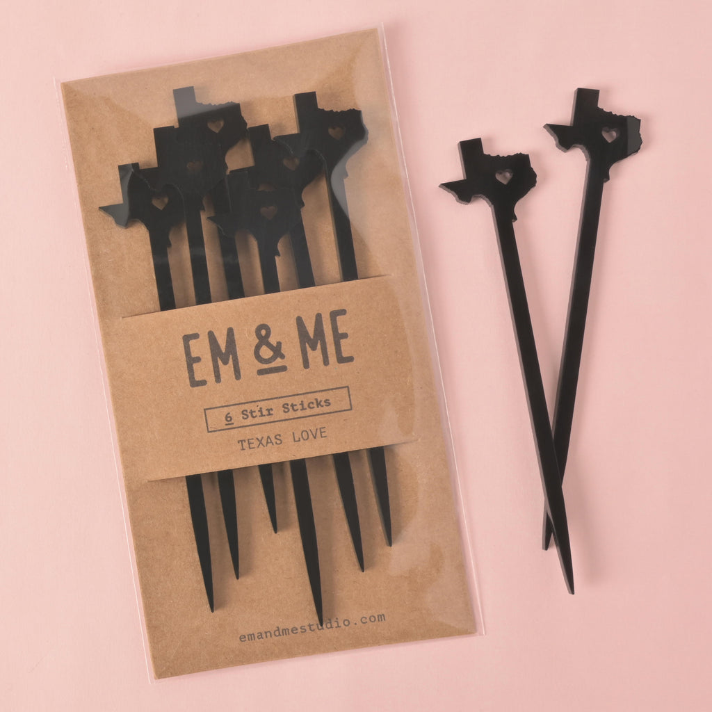 Set of 6 black acrylic drink stir sticks shaped like the state of Texas with heart cutout, packaged by Em & Me Studio on a pink background.