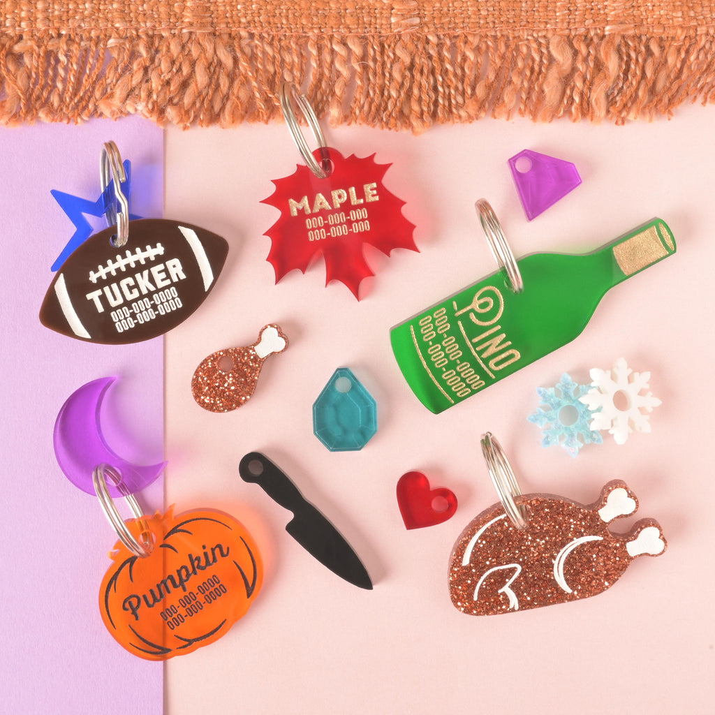 A large collection of personalized pet tags and charms in various shapes and colors scattered on a background of orange knitted fabric, solid purple, and solid pink. The tags include a brown football for "TUCKER," a red maple leaf for "MAPLE," a green wine bottle for "PINO," a neon orange pumpkin for "Pumpkin," a glittery copper turkey, and a variety of small charms like a purple crescent moon, a blue star, a black knife, and a red heart.