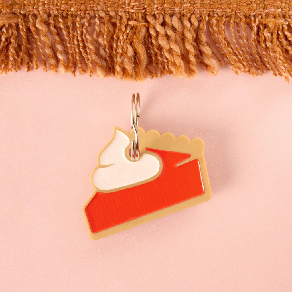 A novelty charm shaped like a slice of pie (pumpkin or sweet potato) made from layered acrylic in tan, red/orange, and white whipped cream. The charm is attached to a split ring and is set against a pink background.