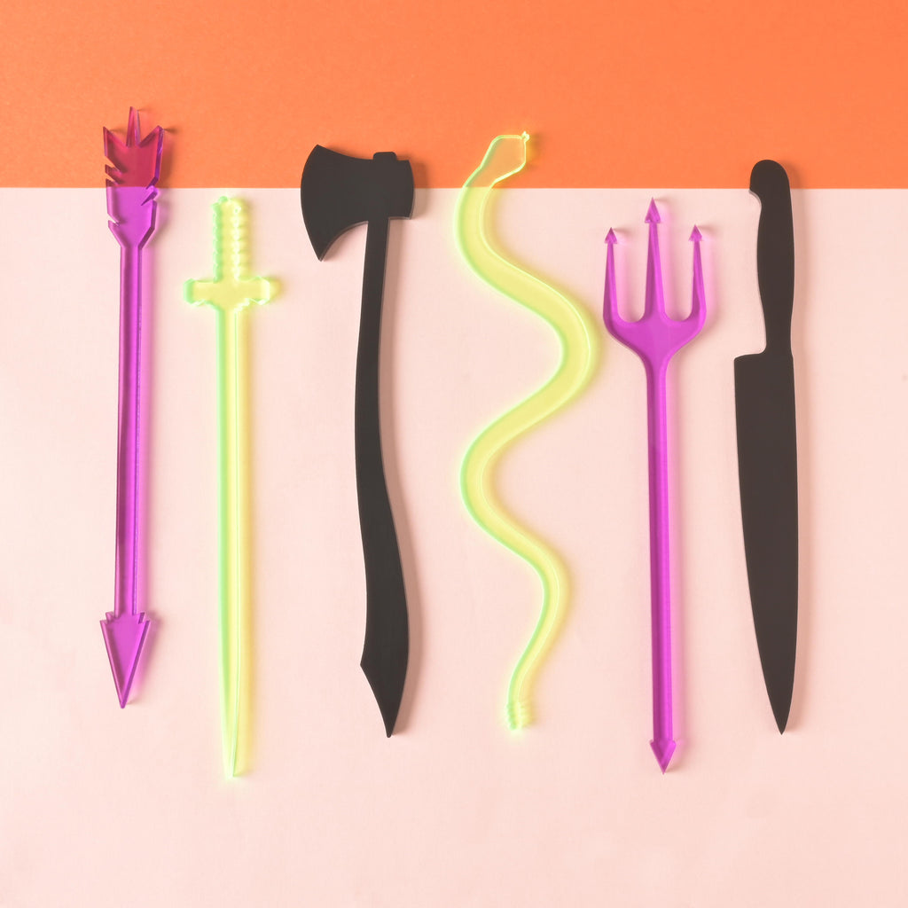 collection of 6 halloween themed stir sticks. purple arrow, fluorescent green sword, black axe, fluorescent green snake, purple pitchfork and black knife on color blocked background