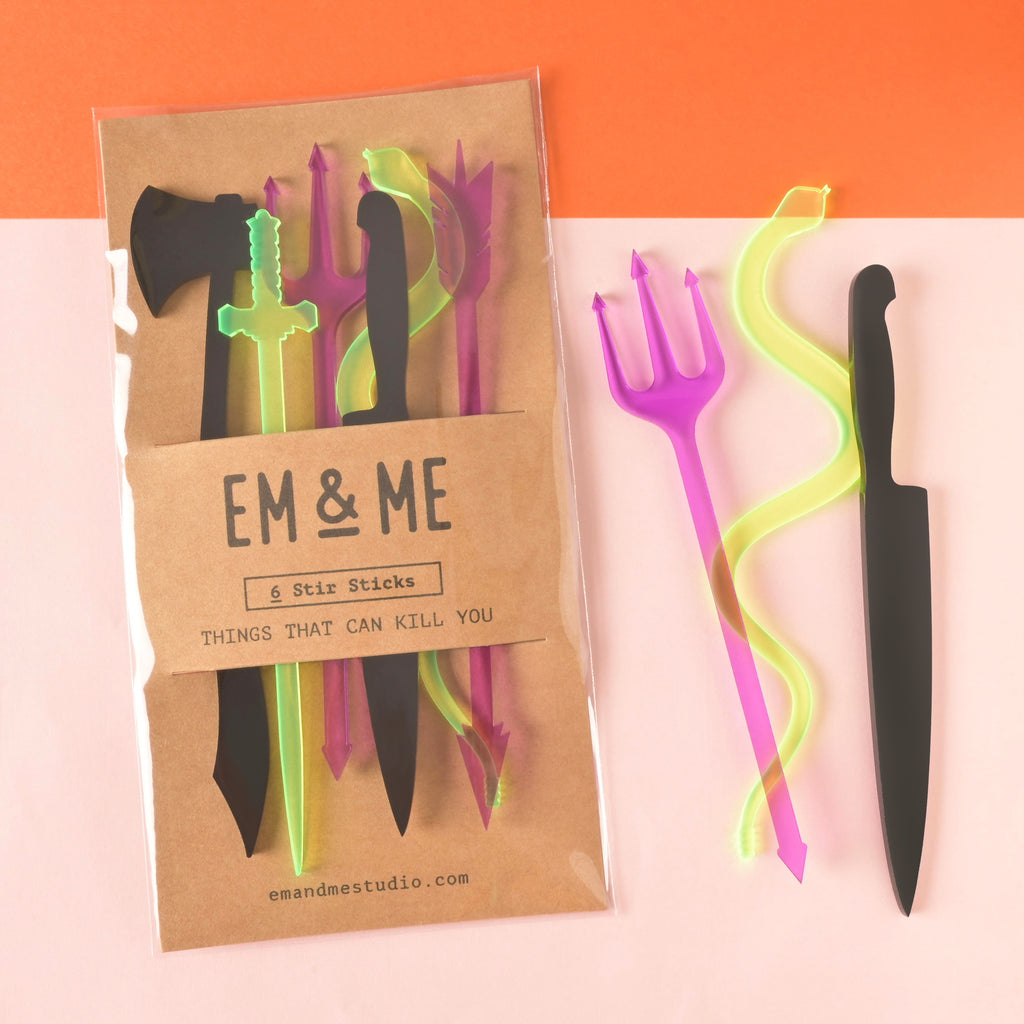 Set of 6 halloween themed stir sticks, shaped like things that can kill you packaged on brown kraft paper with three loose cocktail sticks off to the side on color blocked background