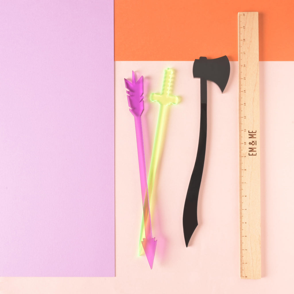 Things that can kill you  set of cocktail stir sticks. purple arrow, fluorescent green sword and black axe next to a wooden ruler on color blocked background