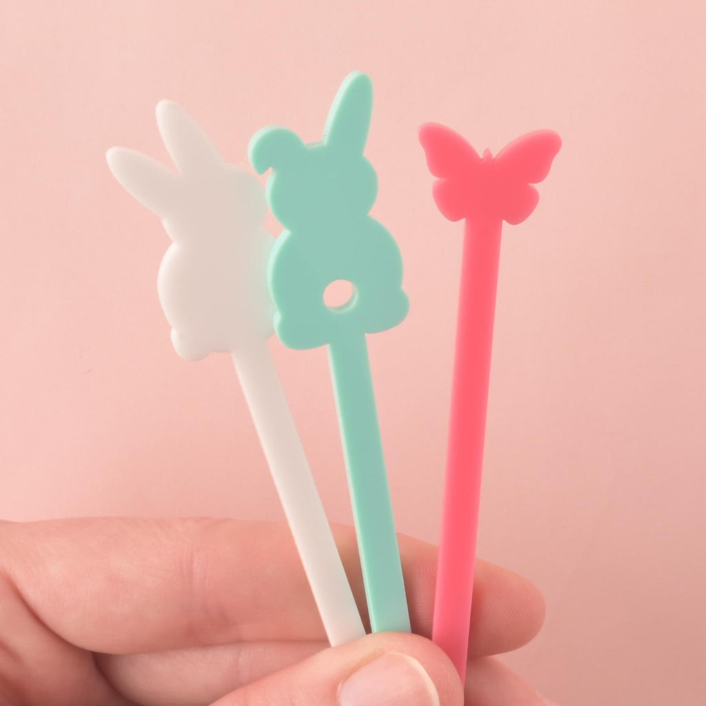 A person holds three Easter-themed cocktail stirrers—a white bunny, a light blue bunny, and a pink butterfly—in their hand. The background is a solid pink color.