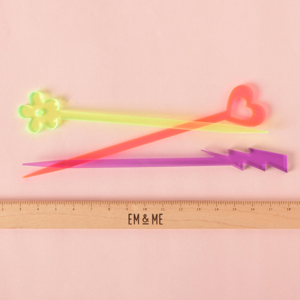 A group of three translucent cocktail stirrers—a fluorescent green  flower, a fluorescent pink heart, and a purple lightning bolt—are shown next to a ruler with the "EM & ME" logo to show their size. The background is a solid pink color.