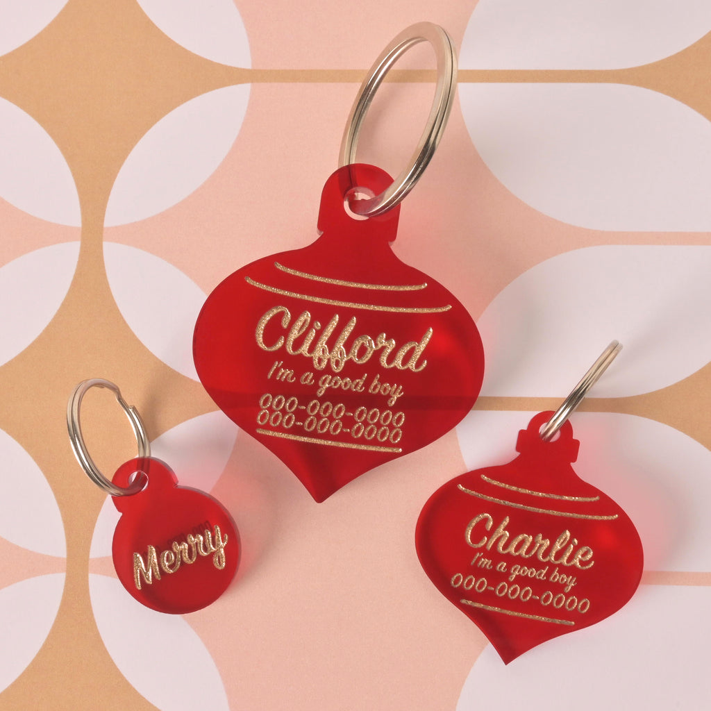 Three red, Christmas ornament-shaped pet ID tags. The tags are engraved with the names "Merry," "Clifford," and "Charlie." The larger tags also include the text "I'm a good boy" and a phone number. They are displayed on a background with a pink and white circular pattern.