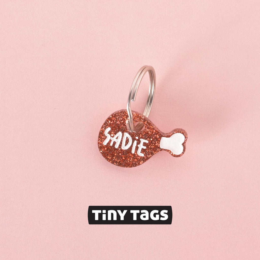 Small glitter drumstick-shaped pet ID tag with the name “Sadie” in white lettering on a pink background, labeled “Tiny Tags.”
