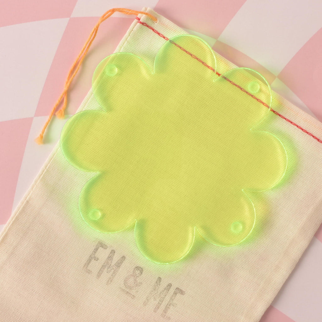 A translucent Fluorescent Green, flower-shaped coaster is displayed on a beige pouch with the "EM & ME" logo. The background is a pink and white checkered pattern.