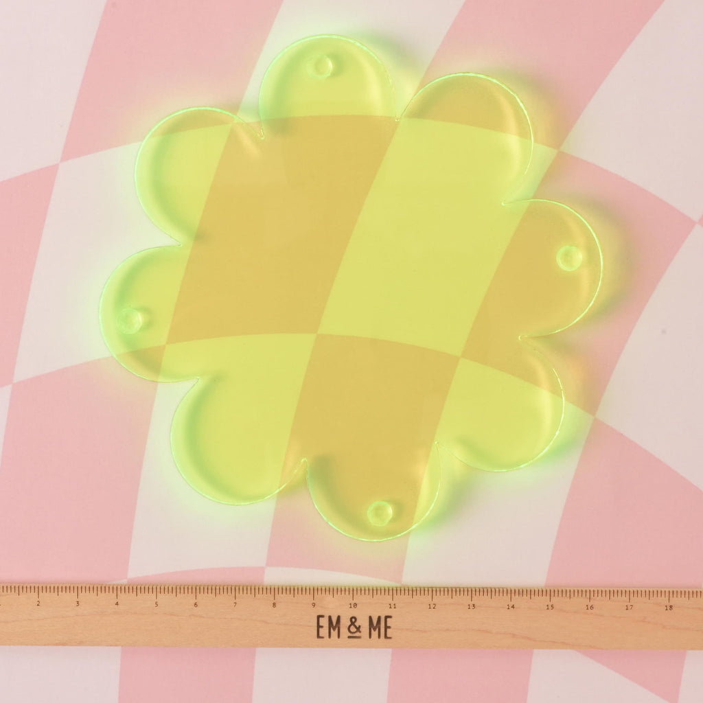 An empty, translucent yellow flower-shaped coaster is displayed on a pink and white checkered surface. A wooden ruler with the "EM & ME" logo rests below the coaster, indicating its size.