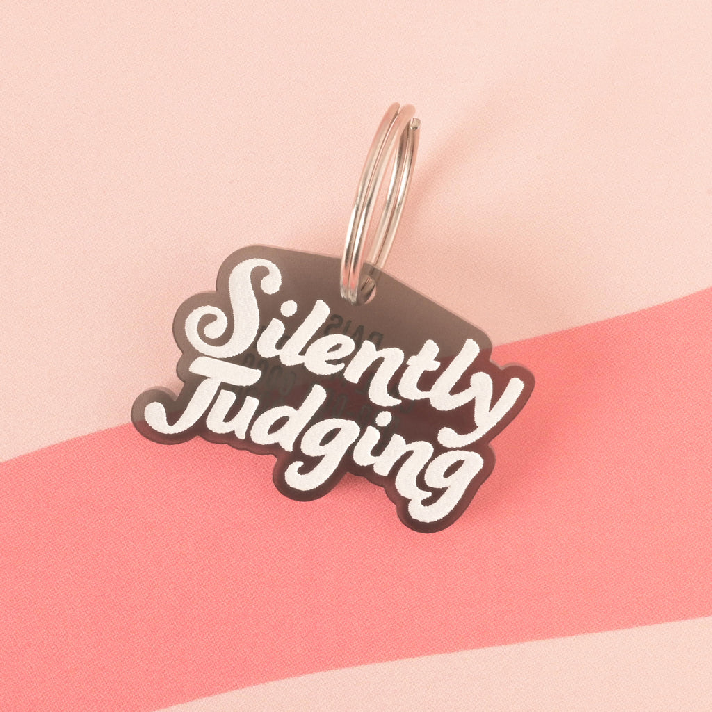 A transparent gray pet tag in the shape of a speech bubble that says "Silently Judging" in a white groovy font. The tag is attached to a silver ring and is resting on a pink background with a wavy design.