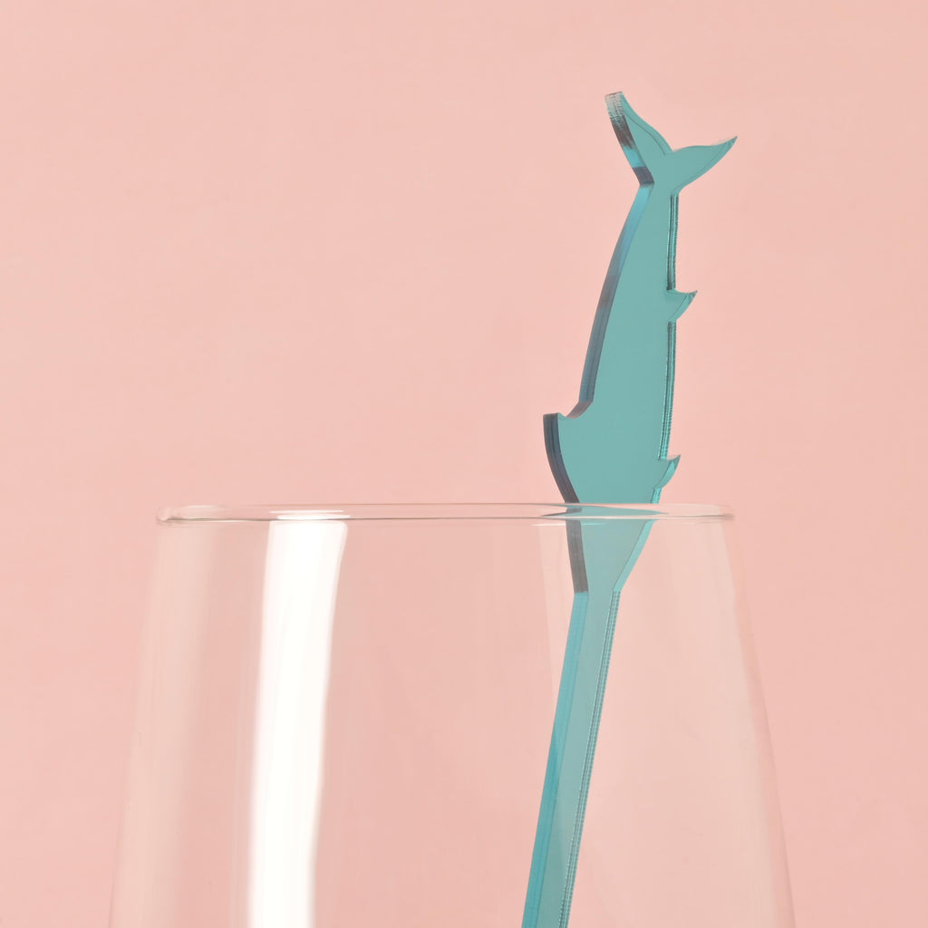 A single blue swordfish-shaped cocktail stirrer is placed inside a clear glass. The background is a solid pink color.