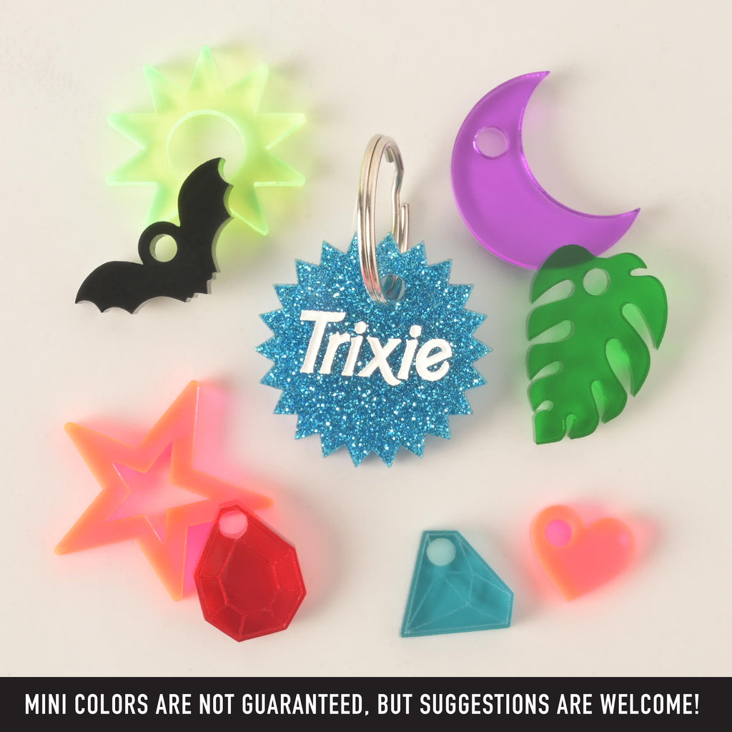 A product photo of a blue glittery, starburst-shaped pet ID tag with the name "Trixie" in the center. Surrounding the main tag are several small, colorful charms: a neon green sun, a black bat, a purple crescent moon, a green monstera leaf, a blue geometric gem, a neon orange heart, a red faceted gem, and a neon orange star. Text at the bottom of the image reads, "MINI COLORS ARE NOT GUARANTEED, BUT SUGGESTIONS ARE WELCOME!"