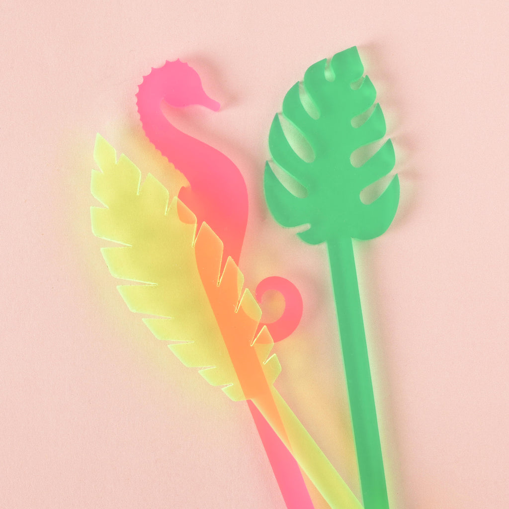 A group of four cocktail stirrers on a solid pink background, including a frosted pink seahorse, a fluorescent green palm frond,  and a green monstera leaf.