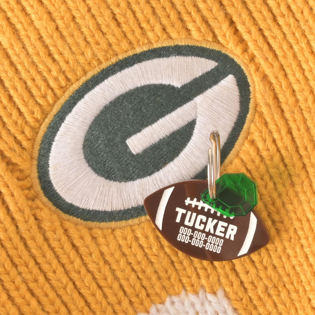 A personalized brown football-shaped pet tag with the name "TUCKER" and a placeholder phone number. The tag has a small green gemstone charm attached. The tag and charm are resting on a yellow and green knitted sweater with a "G" logo, which suggests the Green Bay Packers football team. The tags are connected to a silver ring for a collar.