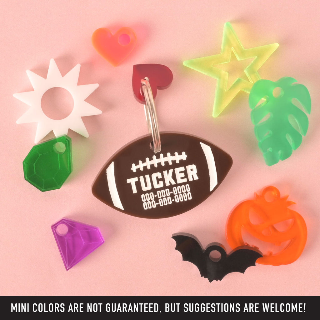 A collection of pet tags and charms scattered on a solid pink background. The central piece is a personalized brown football-shaped pet tag with the name "TUCKER" and a placeholder phone number on it. A small red heart charm is attached. Surrounding it are various charms, including a white sunburst, a green gemstone, a purple gemstone, a neon yellow star, a green monstera leaf, a black bat, and a neon orange carved pumpkin.