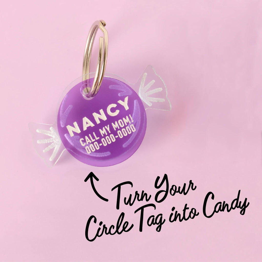 A personalized purple pet ID tag shaped like a wrapped piece of candy, with the name "NANCY" and a placeholder phone number on it. The tag is attached to a silver ring, perfect for a collar, and is displayed on a solid pink background. The image contains text that reads: "Turn Your Circle Tag into Candy."