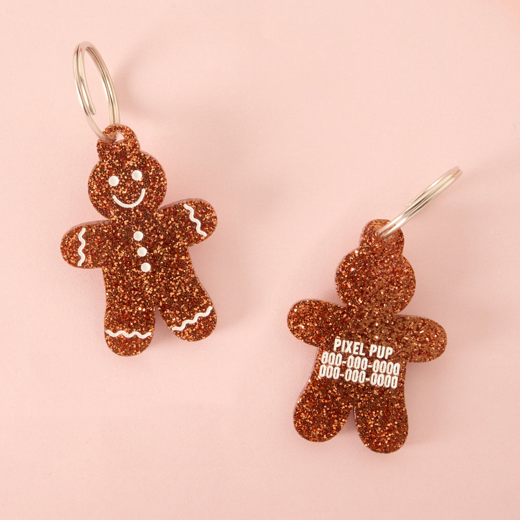 Two views of a gingerbread man-shaped acrylic pet ID tag, featuring a brown glitter finish and white frosting details. The left tag shows the face design, and the right tag shows personalized safety details for 'PIXEL PUP' with phone numbers.