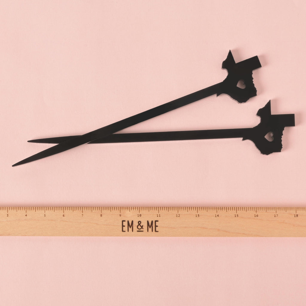 A pair of black cocktail stirrers shaped like the state of Texas, each with a heart cut out in the middle. A ruler with the "EM & ME" logo is placed below the stirrers to show their size. The background is a solid pink color.