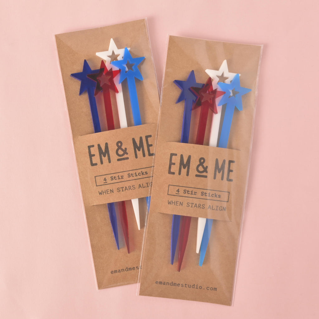Two packaged sets of four red, white, and blue star-shaped cocktail stirrers. Each package is made of brown cardstock with "EM & ME" and "When Stars Align" printed on it. The items are displayed on a solid pink background.