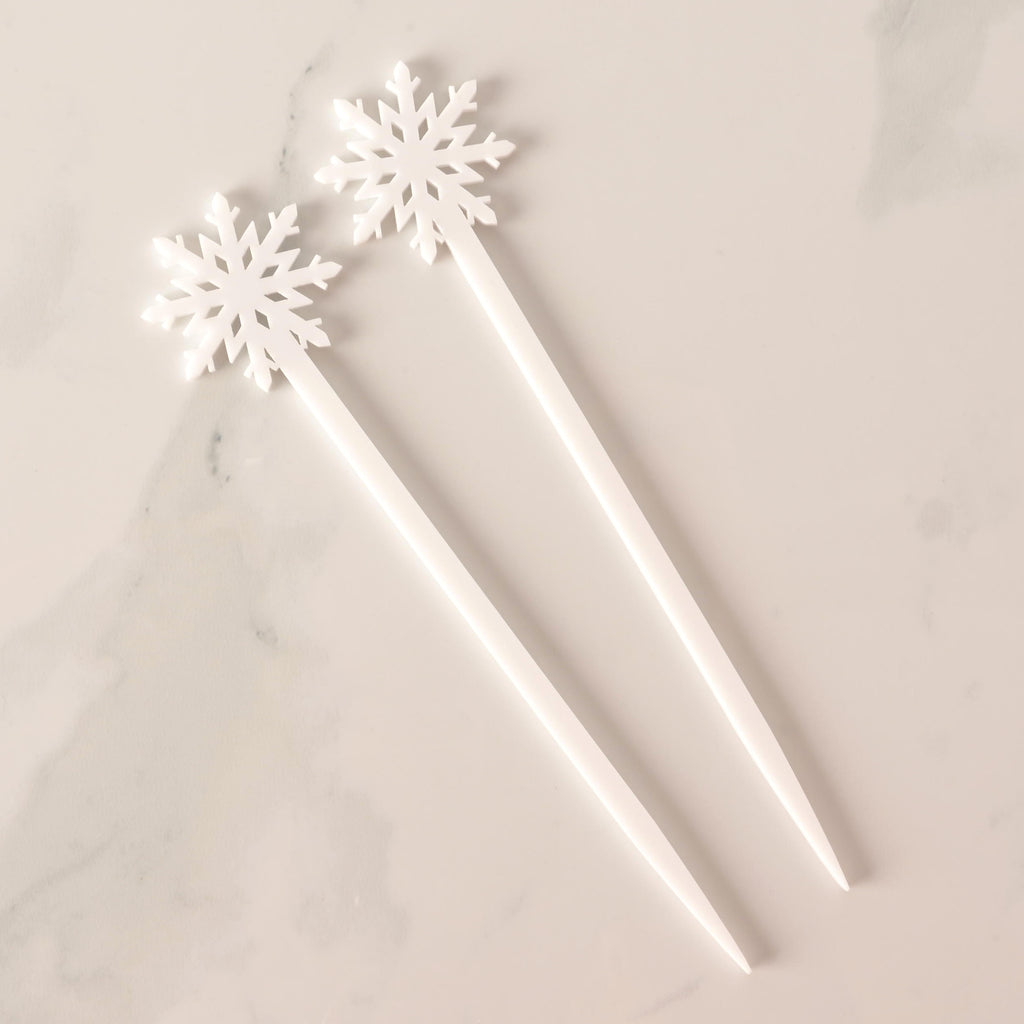 A set of two white snowflake-topped cocktail stirrers, arranged side-by-side on a white marble surface. The stirrers have a clean, solid white appearance.