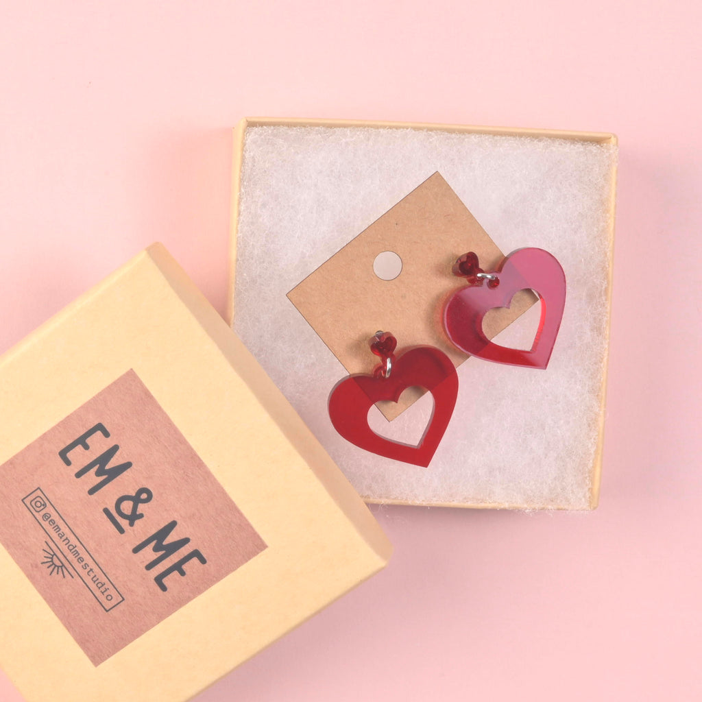 A pair of opaque red acrylic heart-shaped cutout earrings with matching bead accents, displayed on a craft paper earring card inside a small white-lined gift box with an 'EM & ME' branded lid resting alongside.