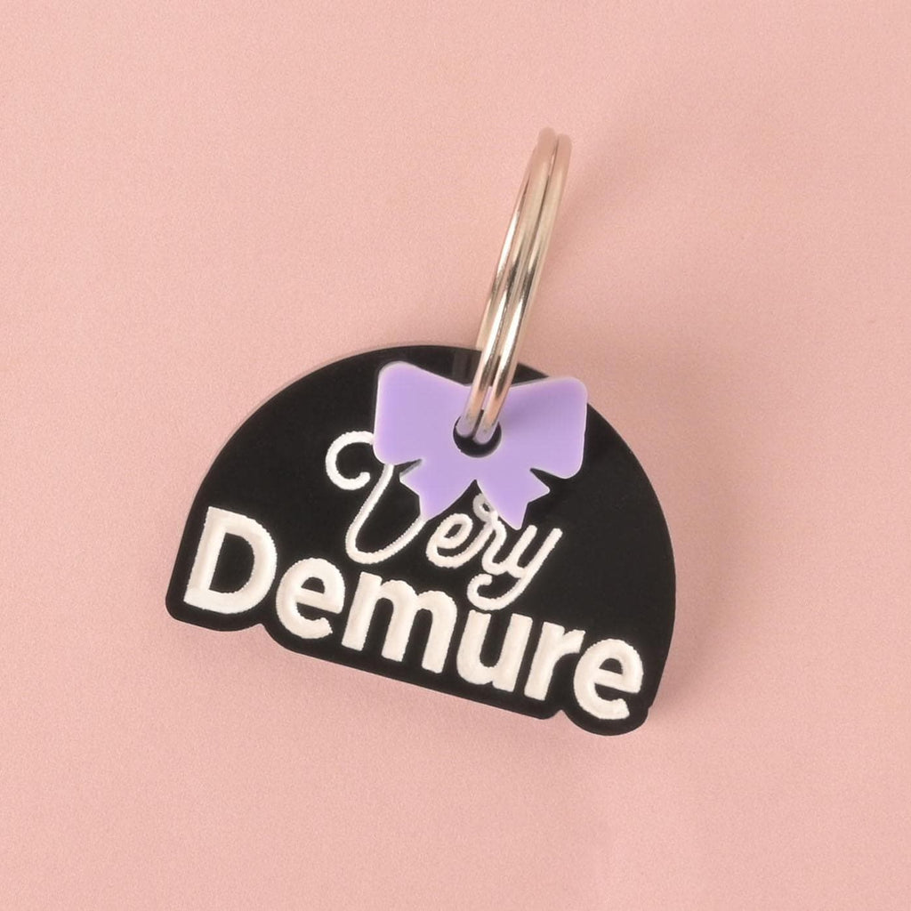 A close-up of a black, half-moon-shaped pet tag with a white engraving that says "Very Demure" in both script and bold font. A small, light purple bow charm is attached to the shiny silver keyring. The tag is shown on a solid pink background.
