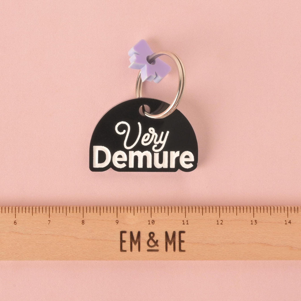 A black, half-moon-shaped pet tag with "Very Demure" engraved in white script and bold font. A small, light purple bow charm is attached to the silver keyring. A wooden ruler with the logo "EM & ME" is visible at the bottom of the photo, which is set on a pink background.