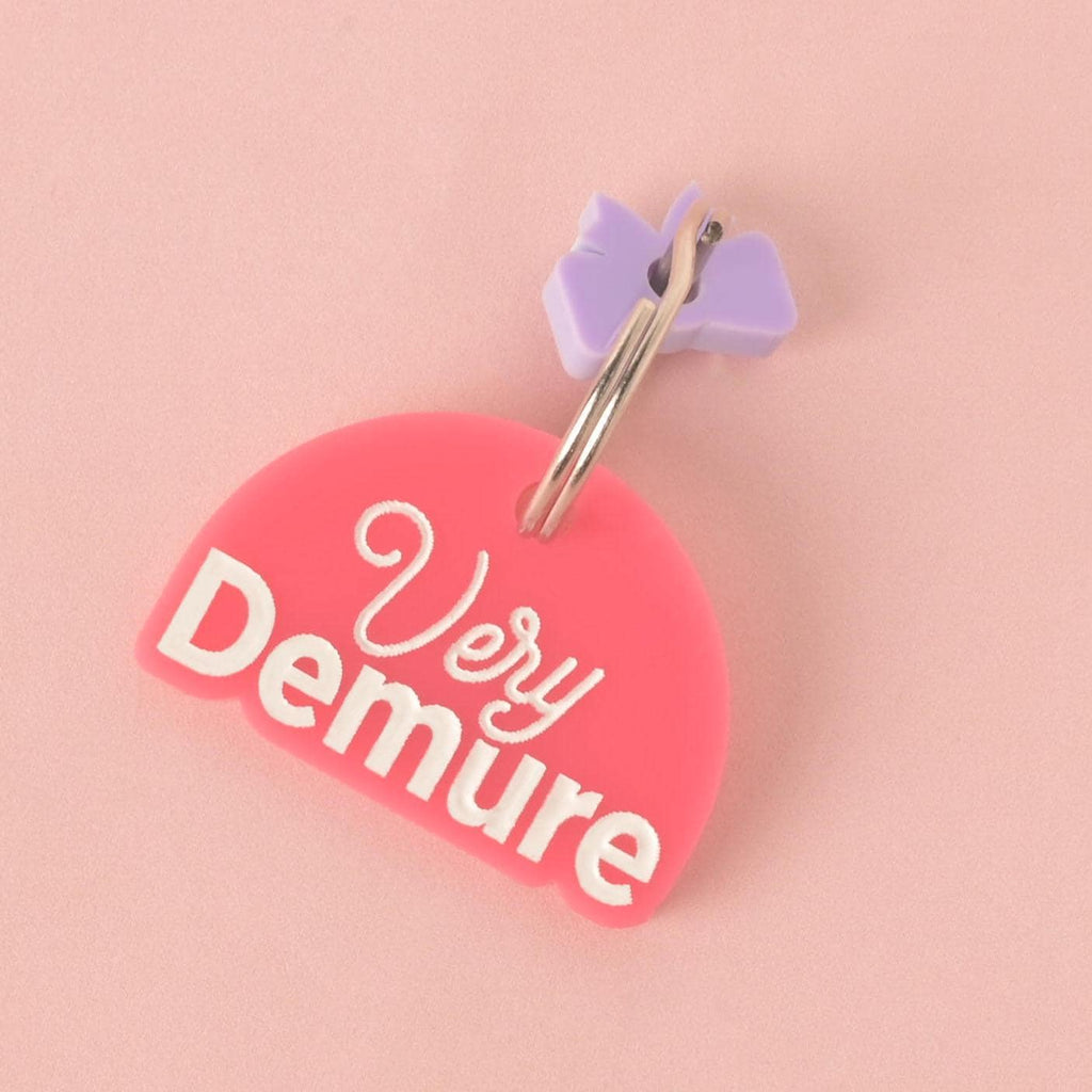 A close-up of a custom, pink, half-moon-shaped pet tag with "Very Demure" engraved in white script and bold font. A small, light purple bow charm is attached to the shiny silver keyring. The tag is shown on a solid pink background.