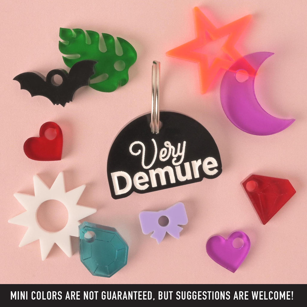 A central black, half-moon-shaped pet tag with a white engraving that says "Very Demure." It is surrounded by a variety of mini charms, including a red heart, a white sun, a blue-green gem, a light purple bow, a red diamond, a purple heart, a purple crescent moon, an orange-red star, a black bat, and a green tropical leaf. The tags and charms are arranged on a pink background. A note at the bottom says, "MINI COLORS ARE NOT GUARANTEED, BUT SUGGESTIONS ARE WELCOME!"