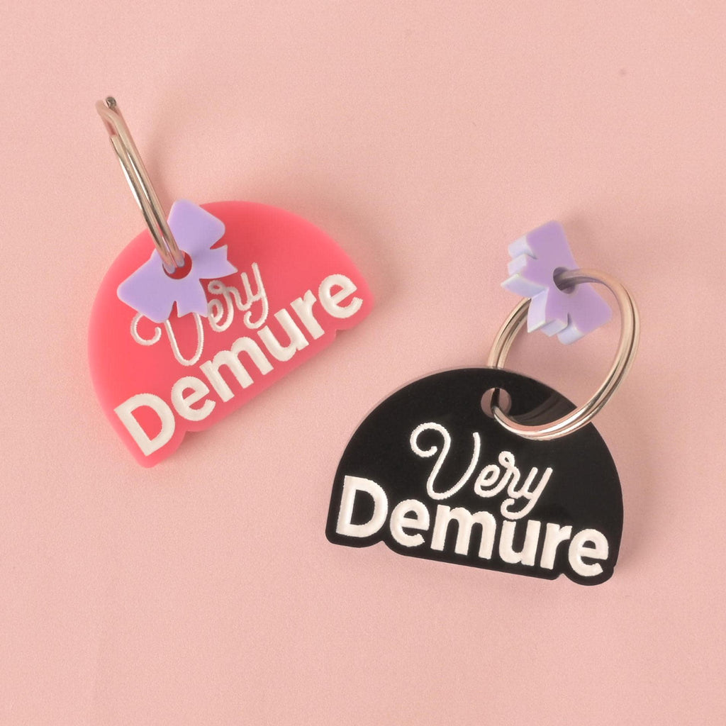 A pair of custom, half-moon-shaped pet ID tags. The tag on the left is pink and the tag on the right is black. Both are engraved with the phrase "Very Demure" in white font and have a small, light purple bow charm attached to a silver keyring. The tags are displayed on a solid pink background.