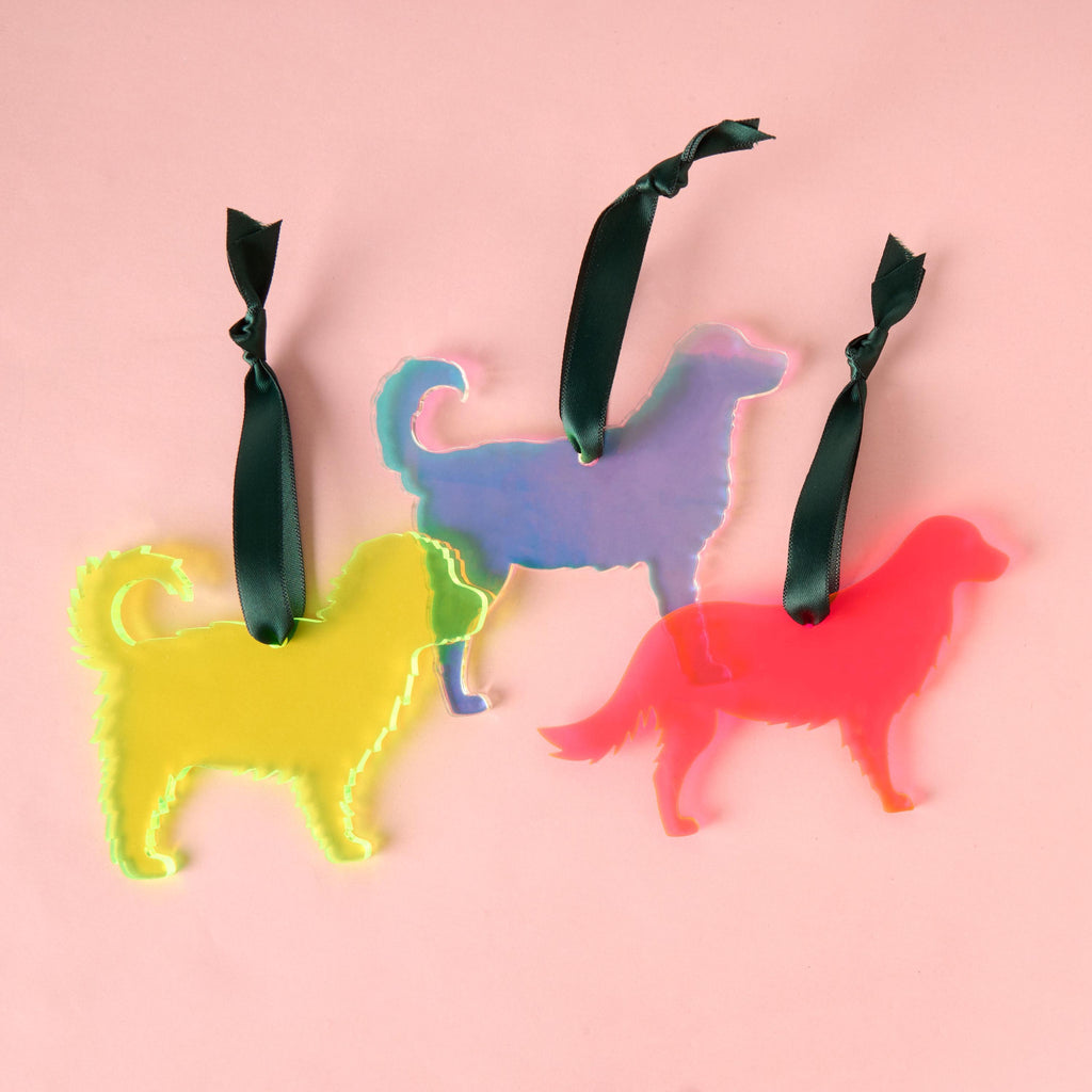 A collection of three laser-cut fluffy dog silhouette ornaments: one fluorescent neon yellow, one vibrant fluorescent red/orange, and one color-shifting iridescent purple/blue. All ornaments are acrylic and hang from dark green satin ribbons.