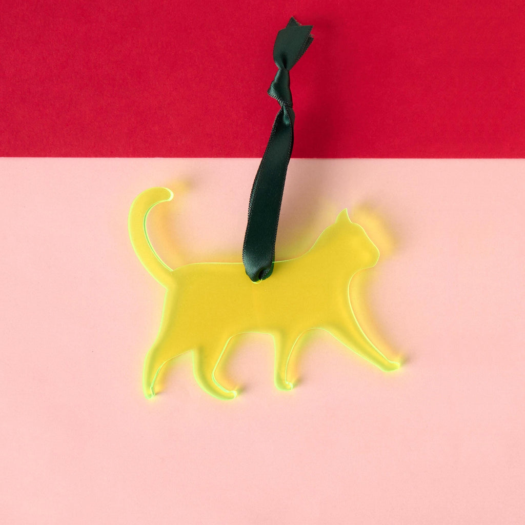 A laser-cut cat silhouette ornament made from vibrant fluorescent neon yellow-green acrylic, featuring a long tail and hanging from a dark green satin ribbon. The ornament is displayed against a two-tone pink and red background.