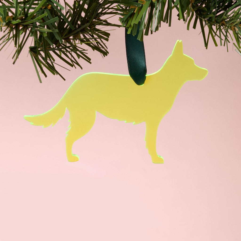 A laser-cut German Shepherd silhouette ornament made from fluorescent neon yellow-green acrylic, hanging by a dark green ribbon from a pine branch, set against a pink background.
