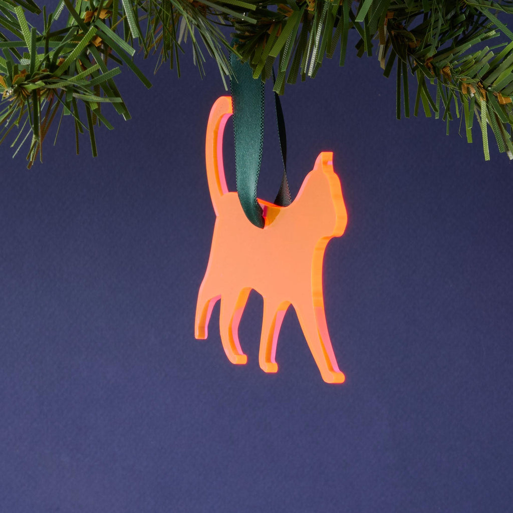 A laser-cut fluorescent pink acrylic cat silhouette ornament, hanging by a dark green ribbon from a pine branch. This vibrant pet décor is set against a dark blue background.