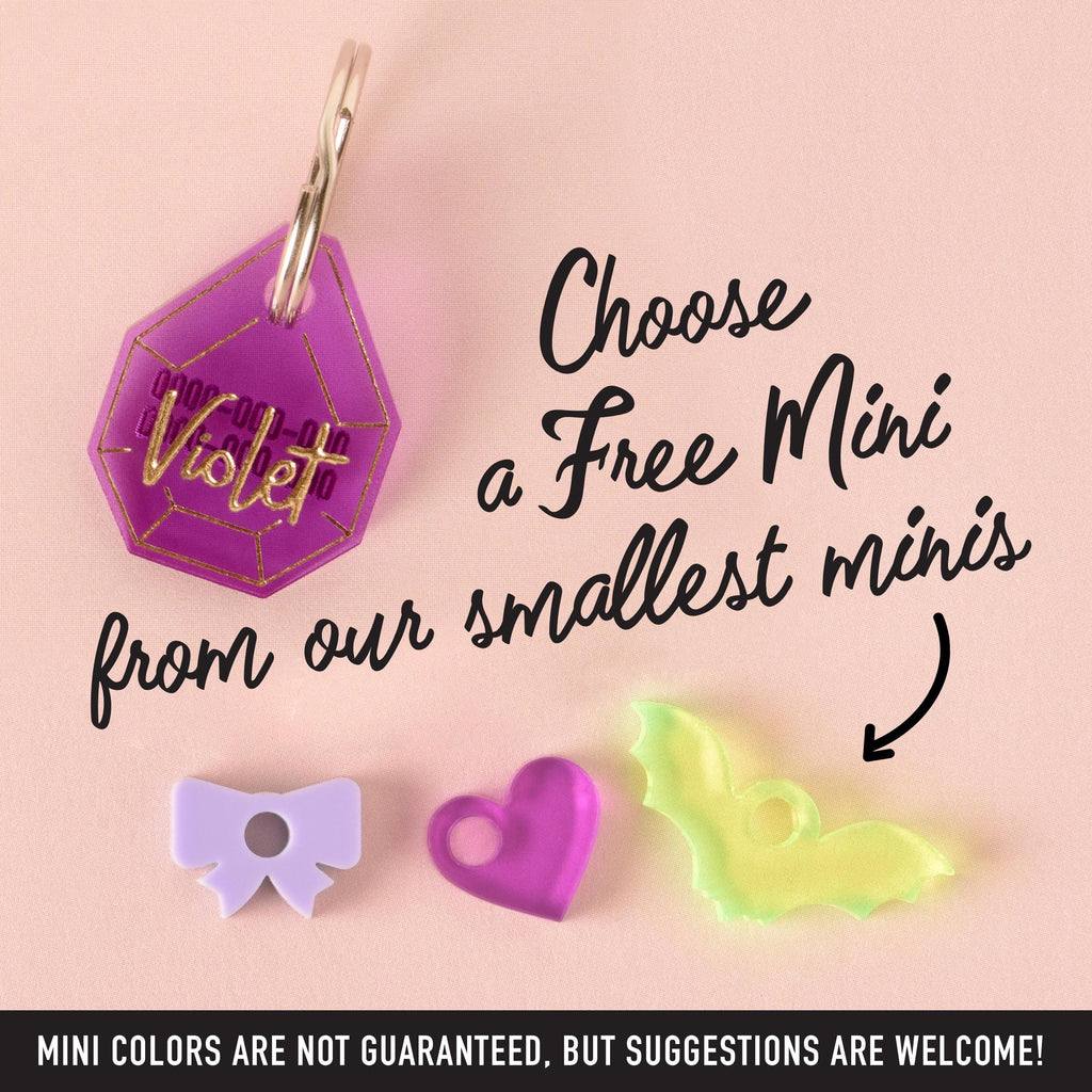 A personalized purple gemstone-shaped pet tag that says "Violet" in a gold-colored script, with a placeholder phone number. The tag is attached to a silver ring and is shown with three small charms: a light purple bow, a pink heart, and a translucent yellow bat. The image also contains the text "Choose a Free Mini from our smallest minis" and "MINI COLORS ARE NOT GUARANTEED, BUT SUGGESTIONS ARE WELCOME!" The items are displayed on a solid pink background.