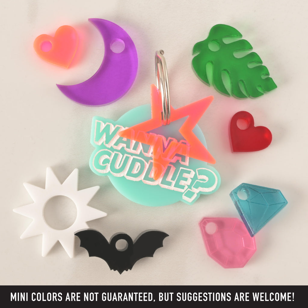 A collection of pet tags and charms scattered on a white surface. The central piece is a personalized aqua-colored tag that says "WANNA CUDDLE?" with a neon orange star charm attached. Surrounding it are various charms, including a purple crescent moon, a green monstera leaf, a red heart, a black bat, a white sun, and two different colored gemstones. The image also contains the text "MINI COLORS ARE NOT GUARANTEED, BUT SUGGESTIONS ARE WELCOME!"