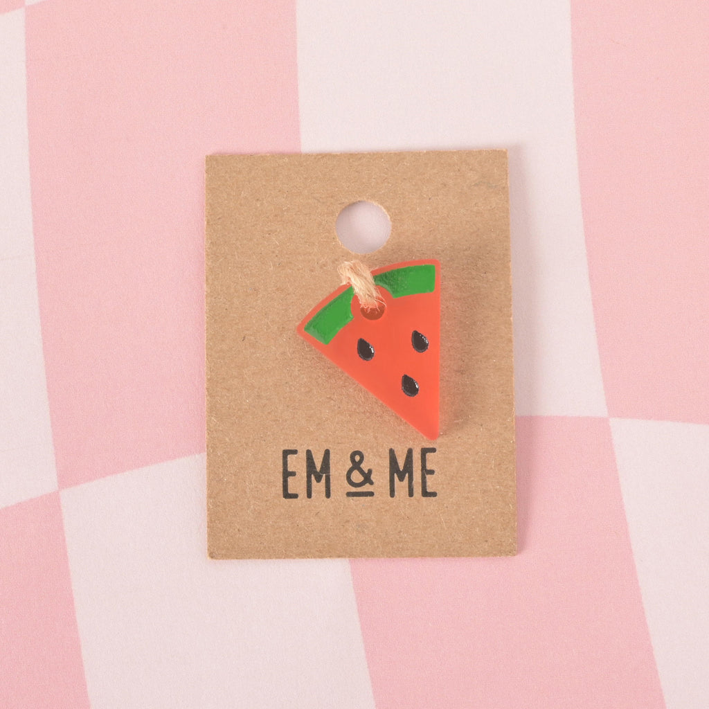 A small, pink and green watermelon slice-shaped pet tag charm with black seeds. The charm is attached with a piece of string to a brown cardboard tag with the logo "EM & ME." The item is displayed on a pink and white checkered background.