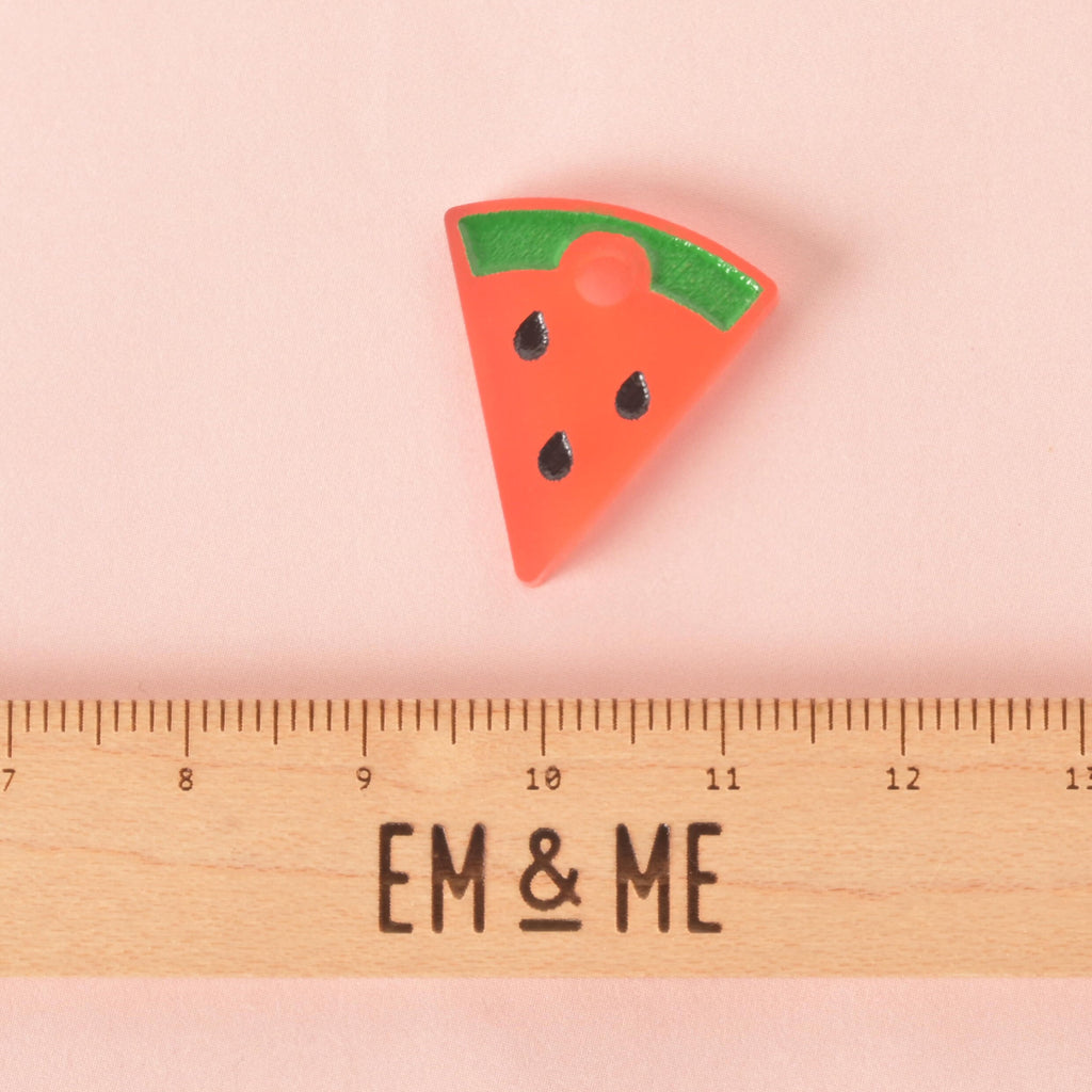 A small, pink and green watermelon slice-shaped pet tag charm with black seeds. The charm is displayed above a wooden ruler with the logo "EM & ME." The background is a solid pink.