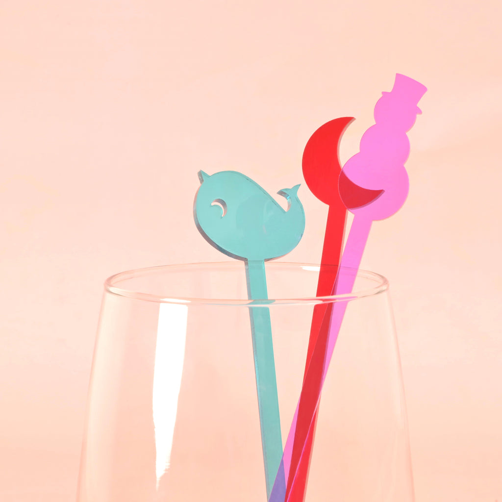 close up of three cocktail stir sticks in a glass. Teal narwhal, red moon and purple snowman in clear glass in front of solid pink background.
