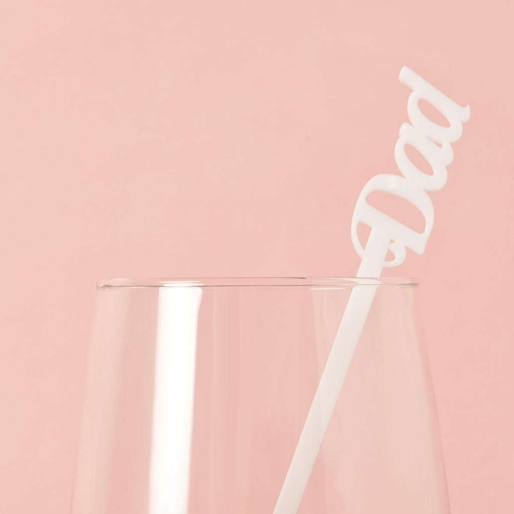 A single white cocktail stirrer with the word "Dad" is placed inside a clear glass. The background is a solid pink color.
