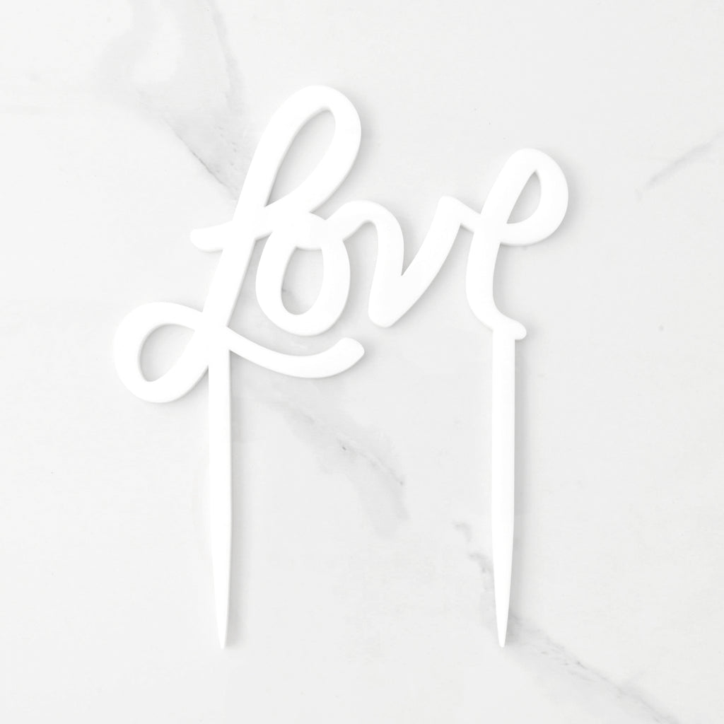 White acrylic cake topper that reads "Love" in a hand drawn script font. Cake topper has two sticks for inserting into cake. shown on marble background